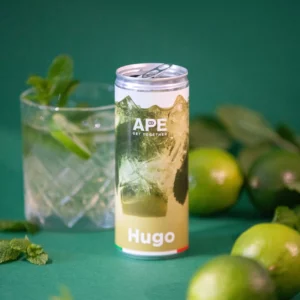 Image of a can of Hugo on a green background. Around the can, there are elements that remind of Hugo: lime, mint, and a glass containing the aperitif.
